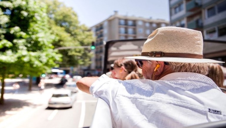 Man wearing a straw hat and earphones enjoying a city tour from an open-top bus.